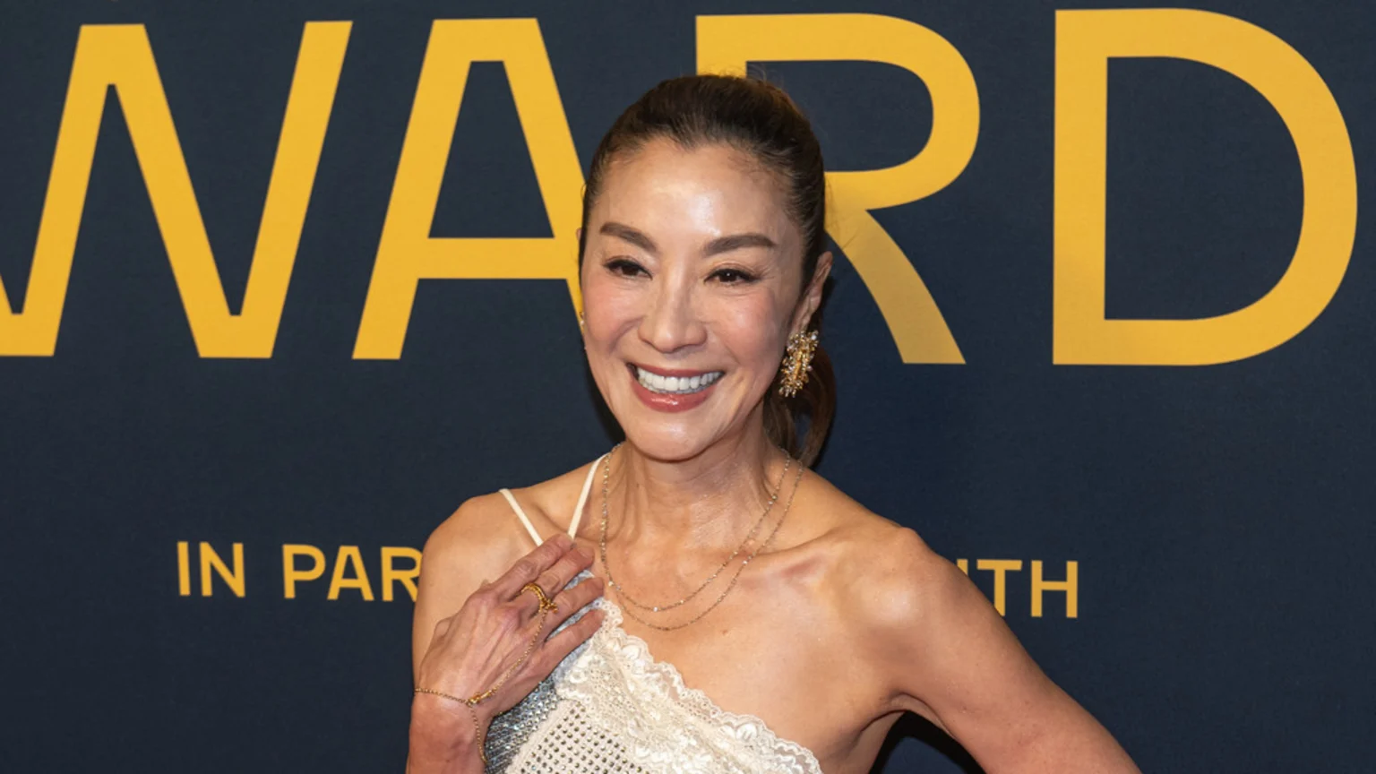 Michelle Yeoh smiles warmly, facing slightly left, in front of a dark blue background with yellow letters spelling "WARD" and "IN PARIS WITH", wearing a white, sparkly, one-shoulder top, layered necklaces, and a large gold earring.