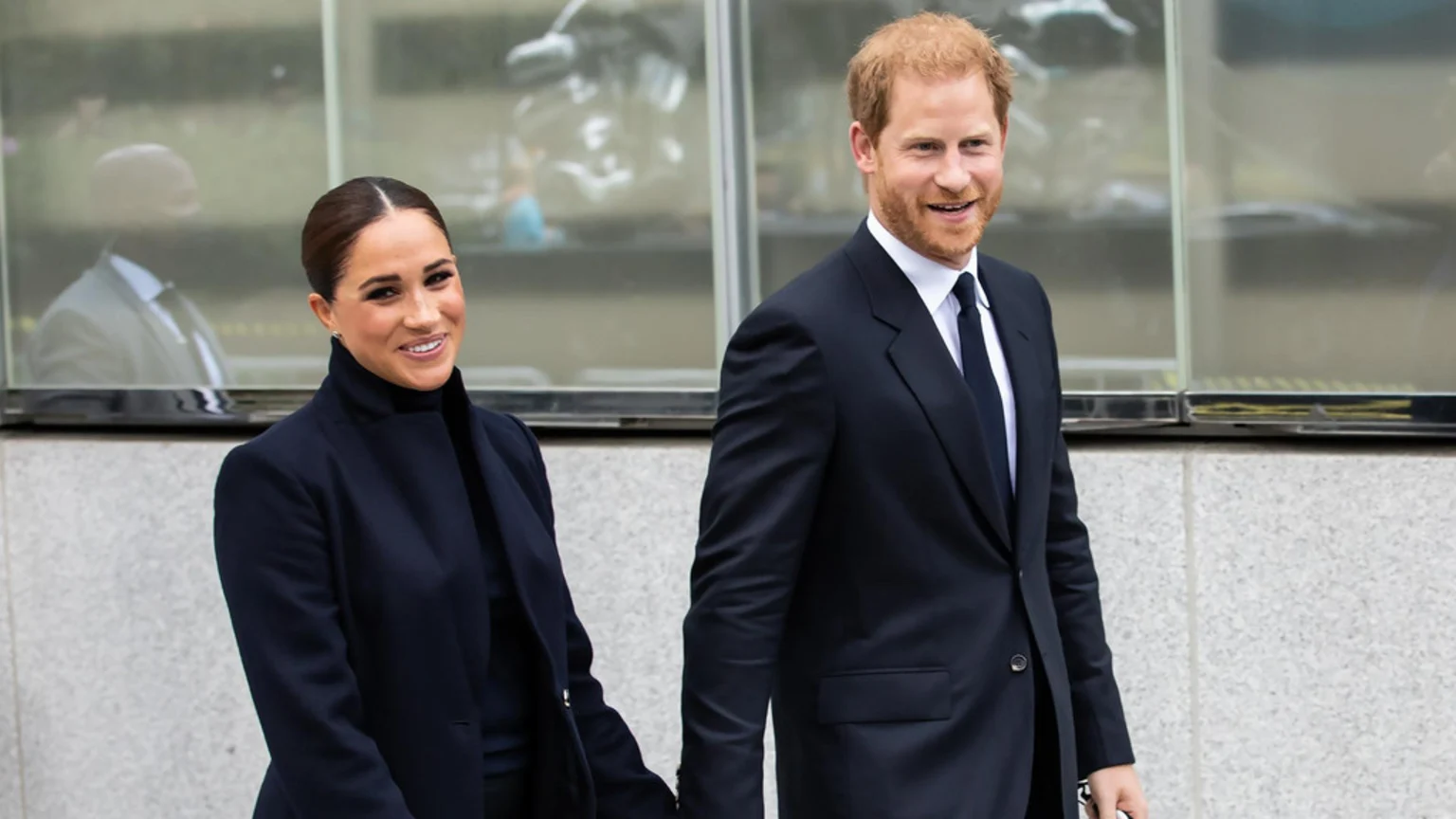 Meghan Markle and Prince Harry smile as they walk forward, Meghan in a dark coat and turtleneck, and Harry in a dark suit, white shirt, and tie, with a blurred building in the background.