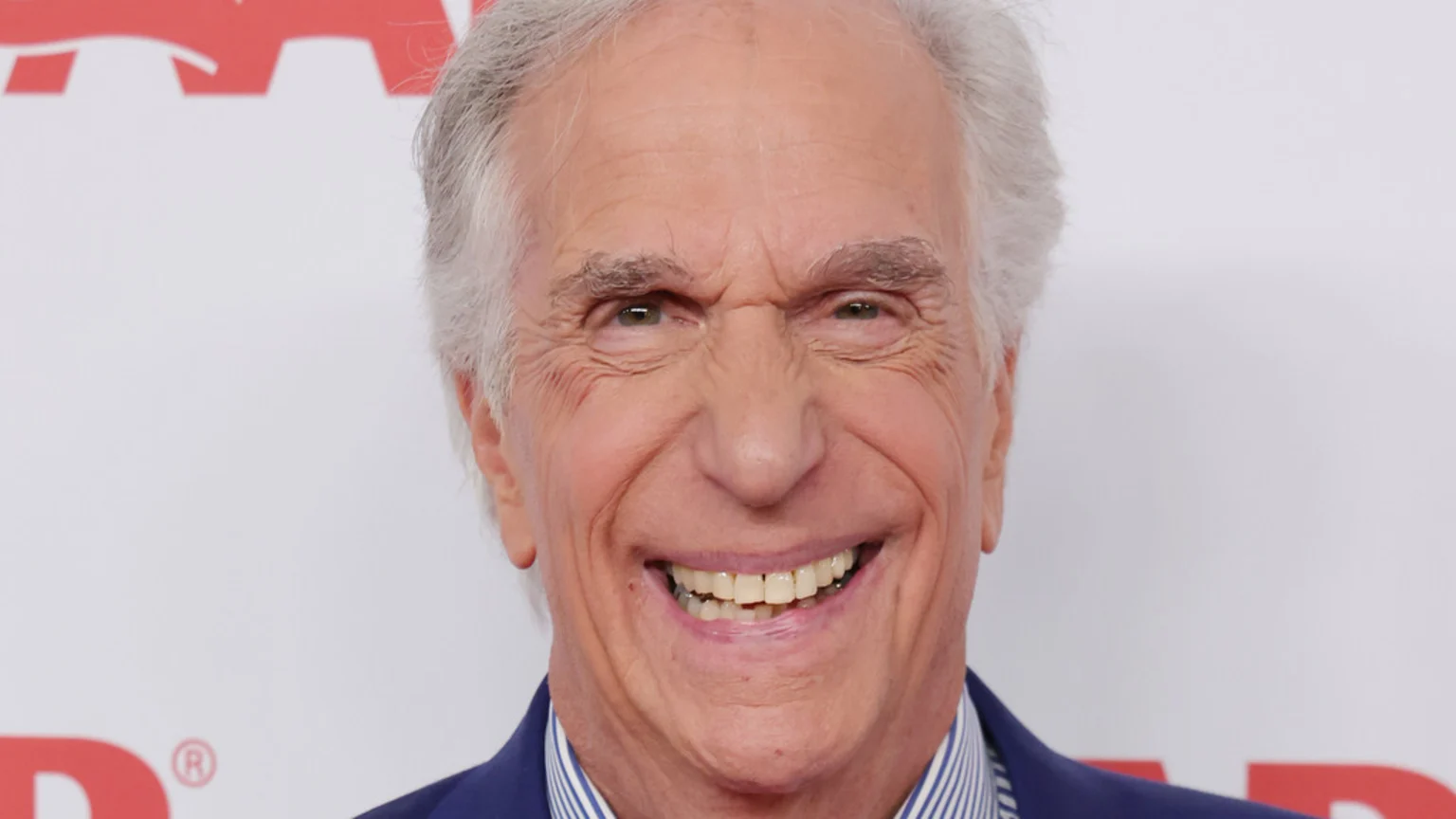 Smiling actor Henry Winkler, with grey hair and a blue suit, smiles widely against a white background with a partial red logo.