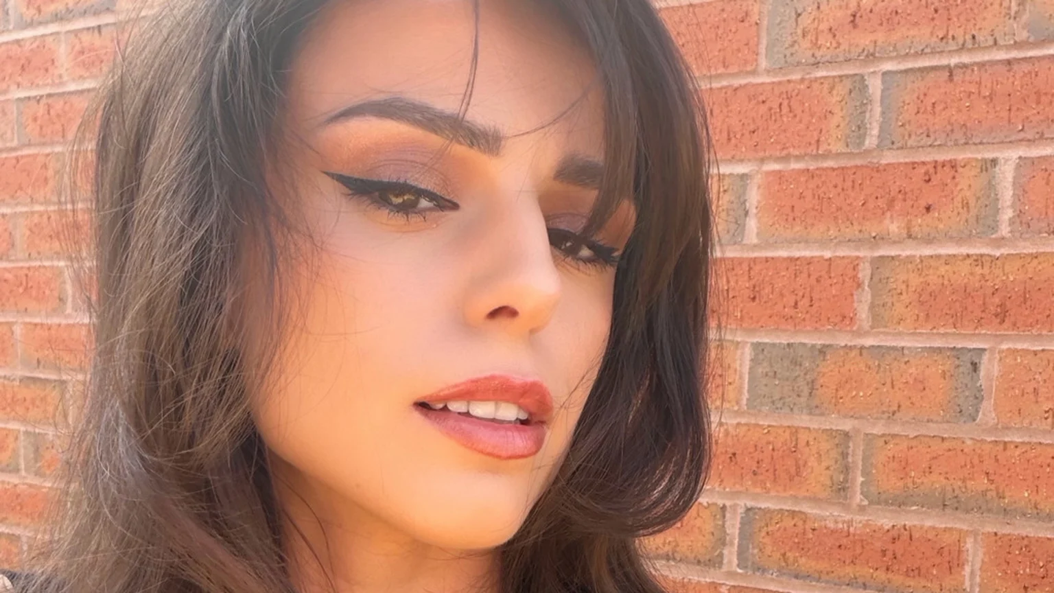 A woman with dark hair and makeup, including winged eyeliner and reddish lipstick, looks towards the camera with a slight smile, illuminated by sunlight against a brick wall.