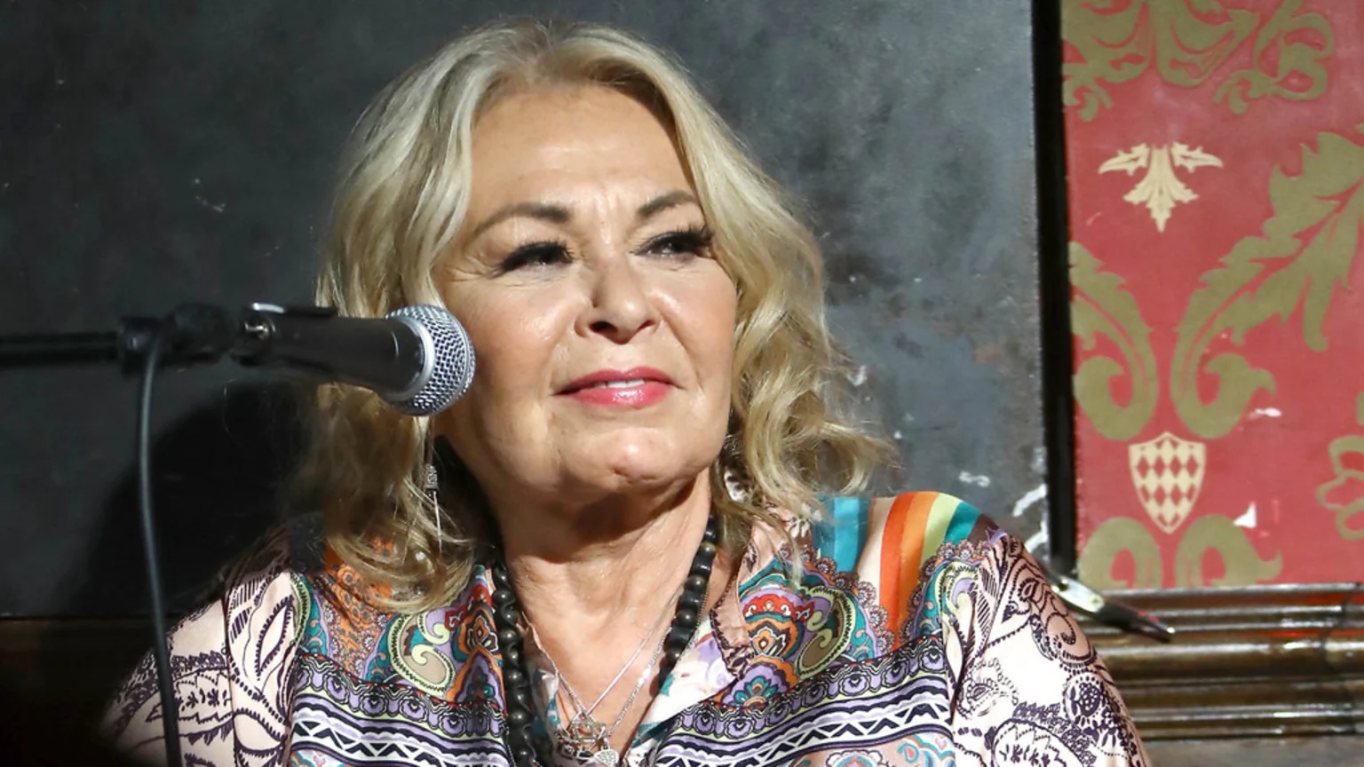 A blonde-haired woman in a colorful patterned shirt with layered necklaces speaks into a microphone, against a dark and red-patterned background.