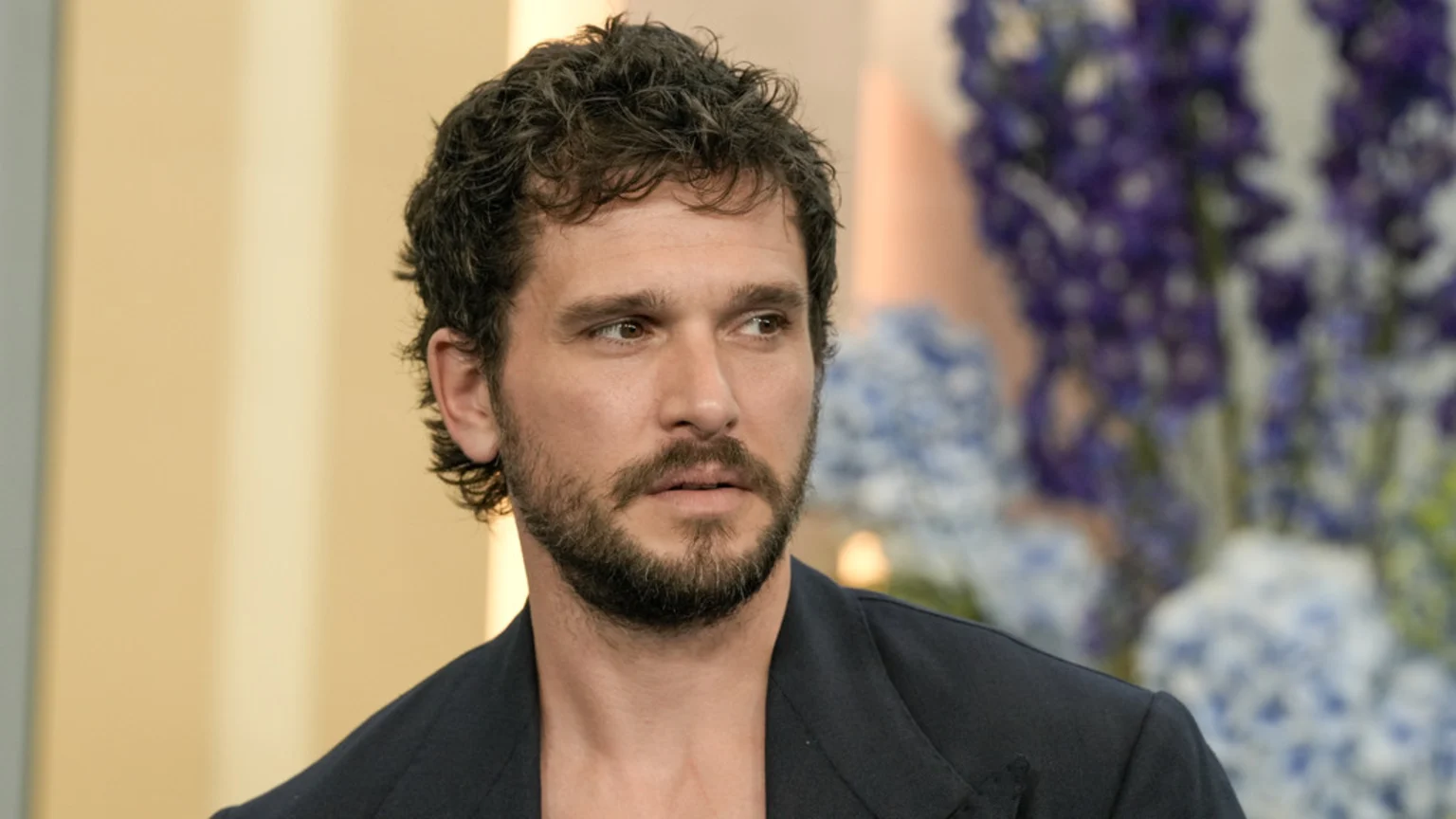 A man with dark curly hair and a beard, wearing a dark open-necked jacket, looks pensively to the side with a blurred background of yellow and purple flowers.