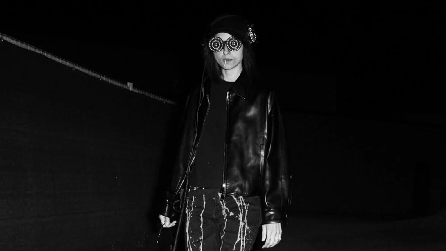 A person in a black cap, headphones, and unique round, studded sunglasses holds a lollipop stick in their mouth, wearing a black leather jacket and paint-splattered pants against a dark, monochromatic background.