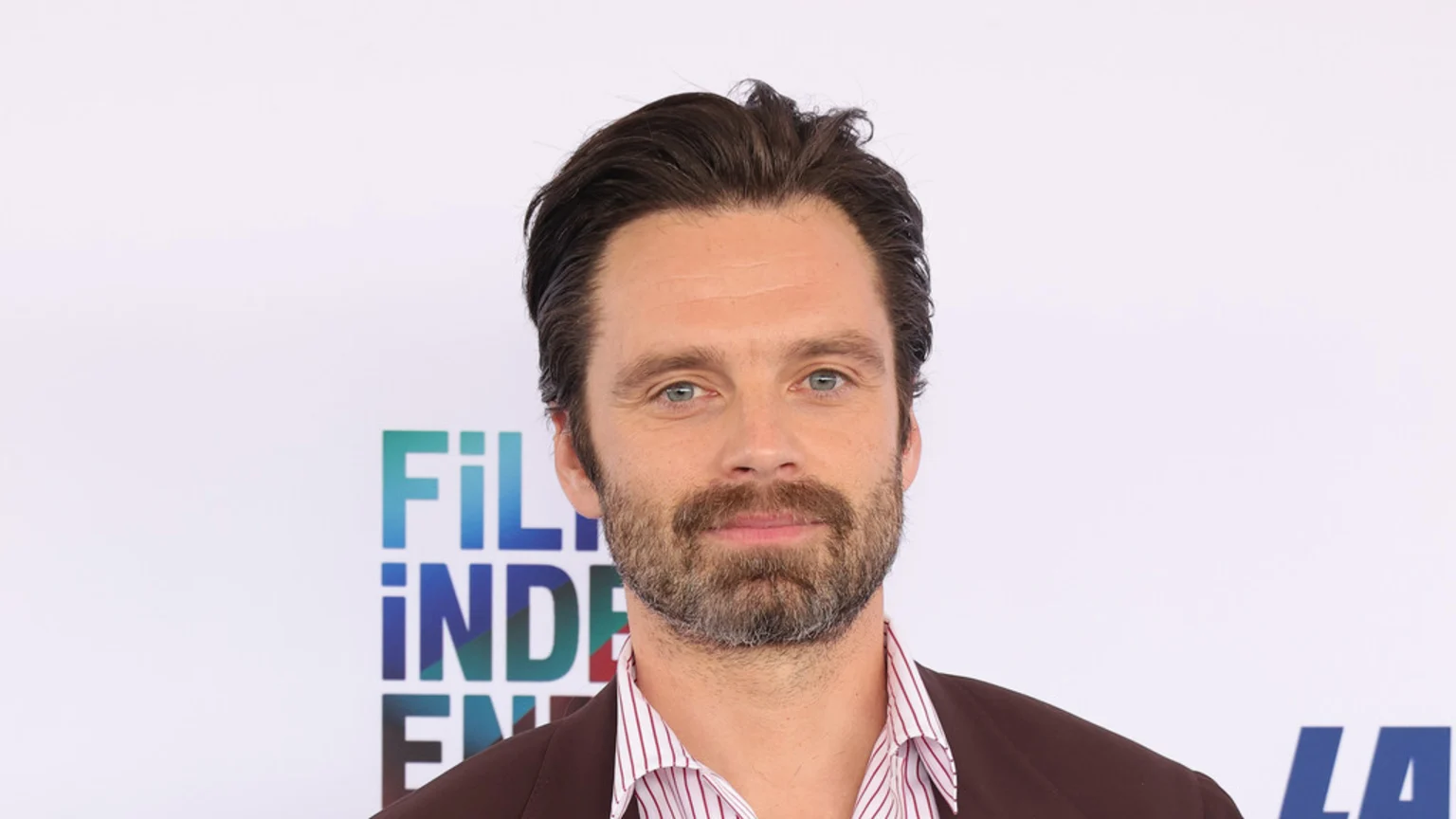Actor Sebastian Stan, with dark hair and a beard, wearing a brown jacket and striped shirt, looks directly at the camera against a white background with blurred "FILM INDEPENDENT" text.