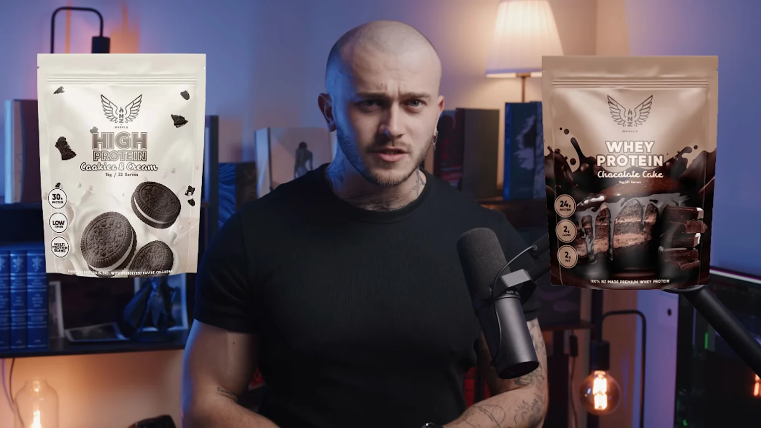 A bald man with tattoos speaks into a microphone, positioned between two floating product bags for "HIGH PROTEIN Cookies & Cream" and "WHEY PROTEIN Chocolate Cake."