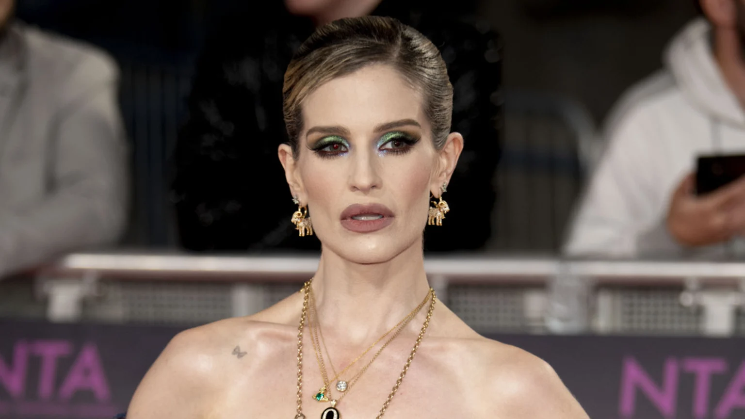 Kelly Osbourne on a red carpet, wearing dramatic green and blue eyeshadow, light brown lipstick, layered gold necklaces, and elephant earrings, with a butterfly tattoo on her shoulder.