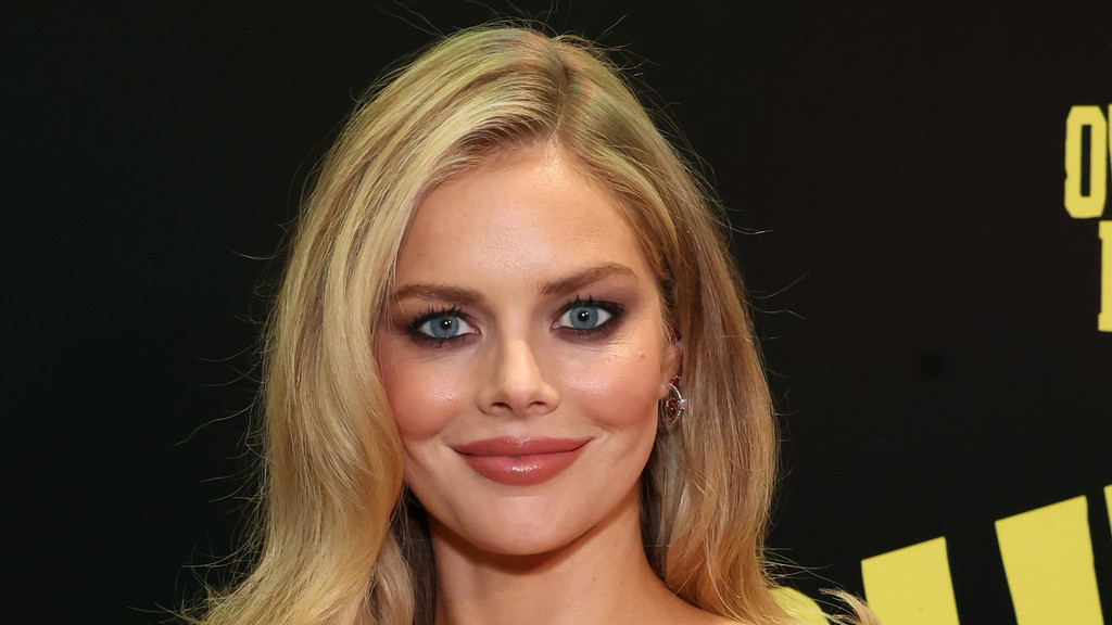 A close-up portrait of actress Samara Weaving smiling, featuring her long blonde hair, bright blue eyes with dramatic makeup, and glossy lips against a dark background.