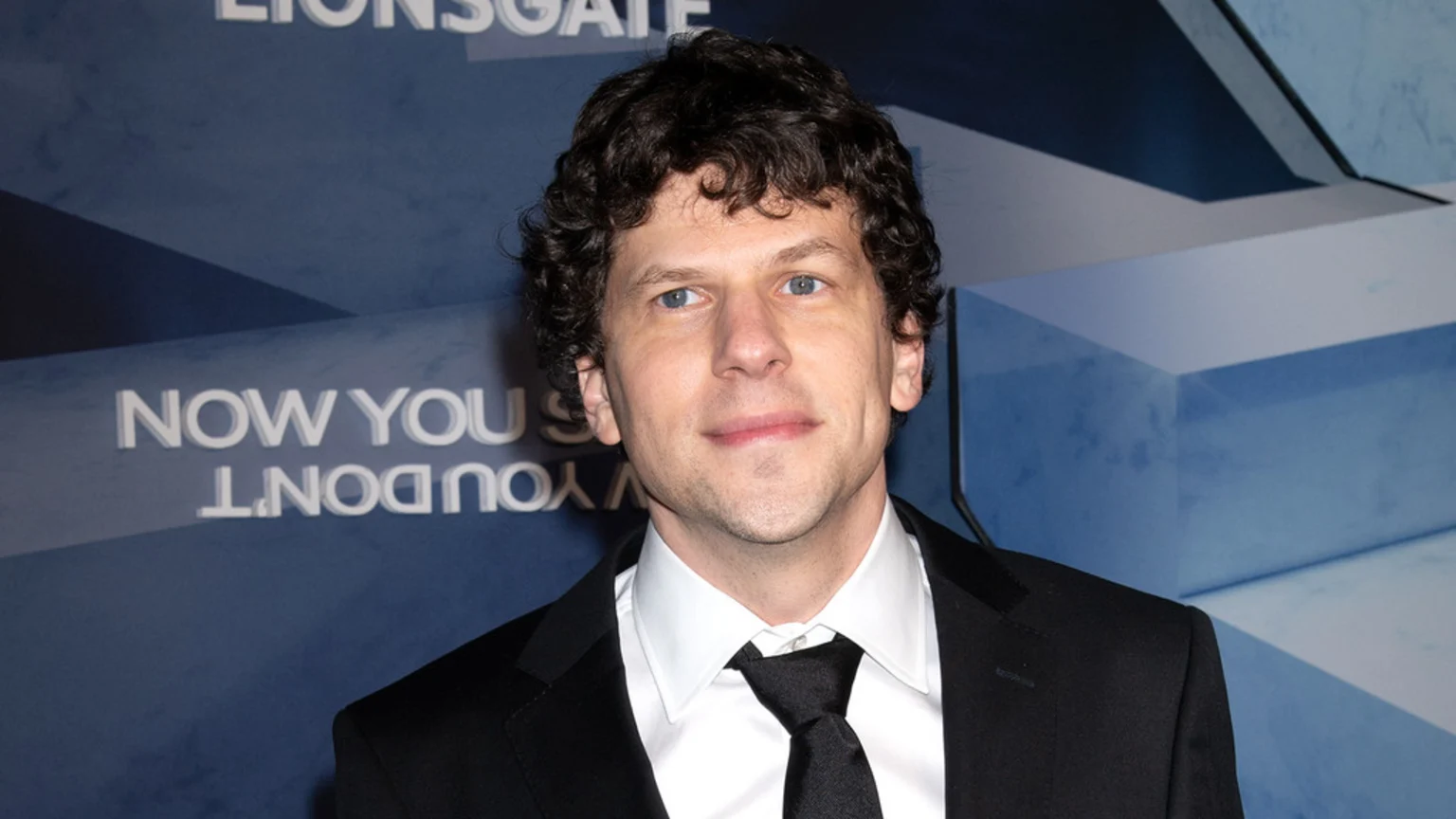 Jesse Eisenberg, with curly dark hair and blue eyes, poses in a black suit and tie against a blue background with "LIONSGATE" visible.