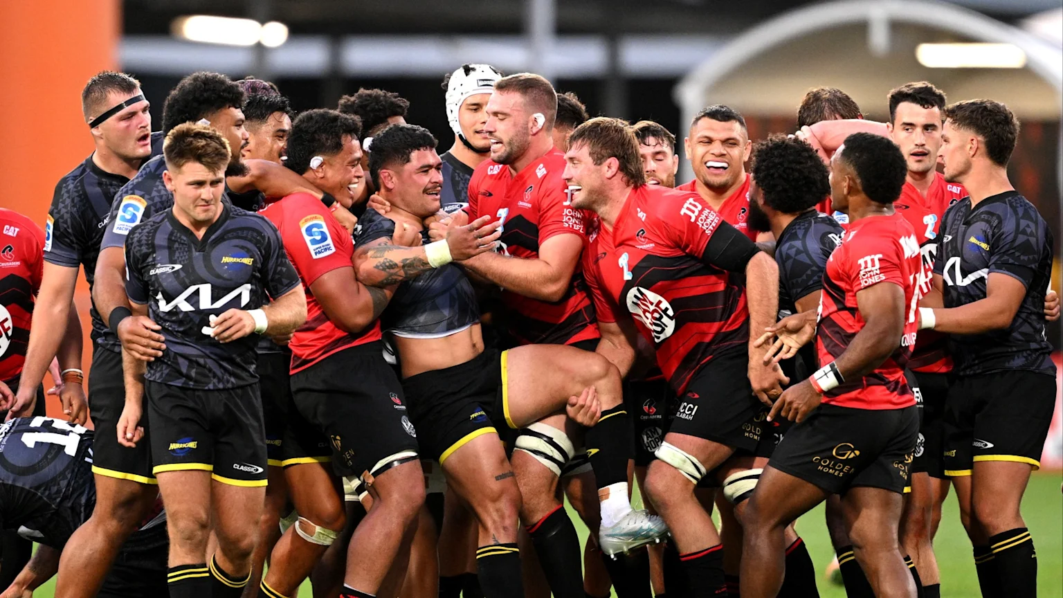 Rugby players from the Crusaders (red and black jerseys) and Hurricanes (dark grey and yellow jerseys) engage in a close huddle on the field, with some smiling and others looking focused.