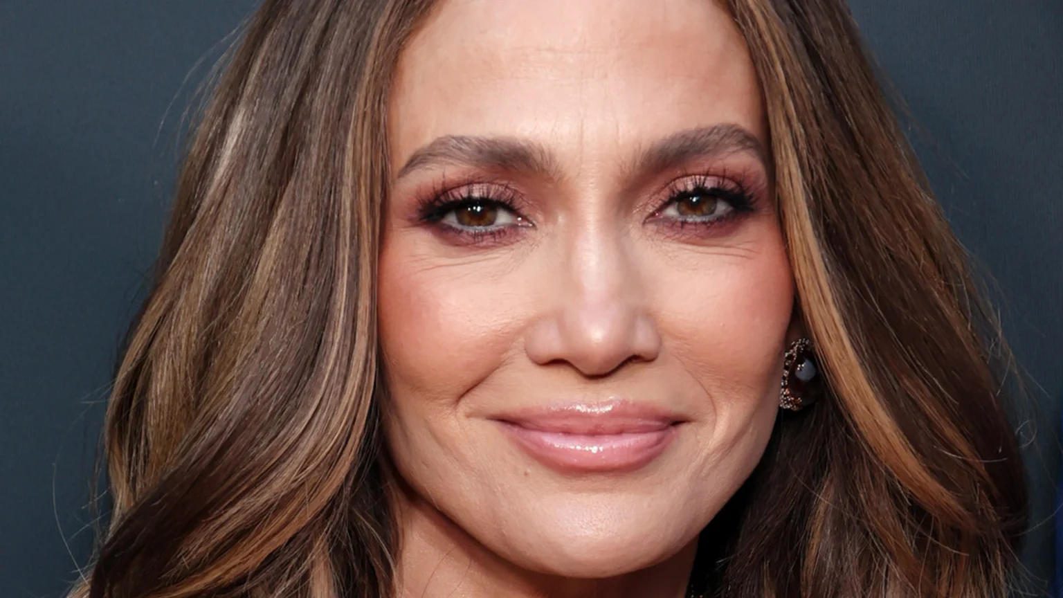 Close-up of Jennifer Lopez smiling, with brown wavy hair, pink eyeshadow, glossy pink lips, and an earring, against a dark background.