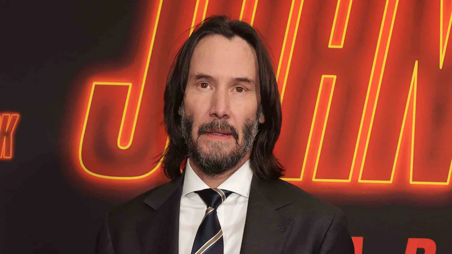 Keanu Reeves, with long dark hair and a beard, wearing a suit and tie, stands in front of a red and black background with neon "JOHN WICK" lettering.