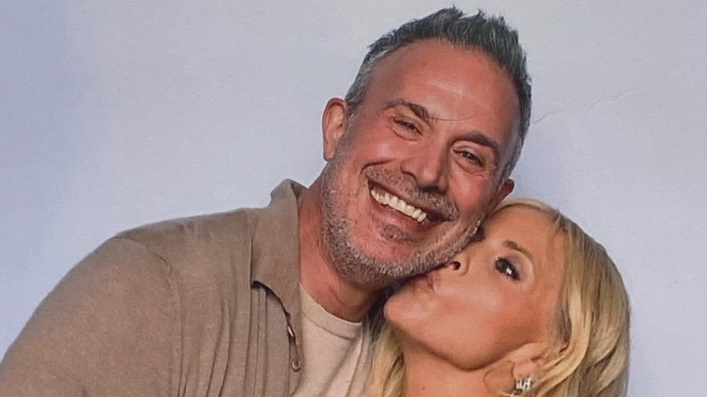 A happy man with grey hair and beard smiles widely as a blonde woman affectionately kisses his cheek.