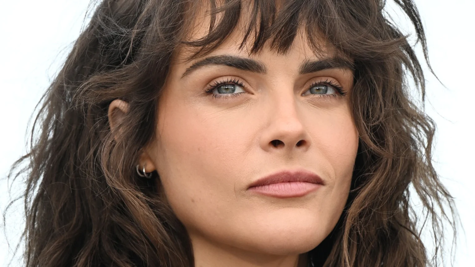 A headshot of a woman with long, dark, wavy hair and bangs. She has fair skin, blue-green eyes, and wears muted pink lipstick and small hoop earrings.