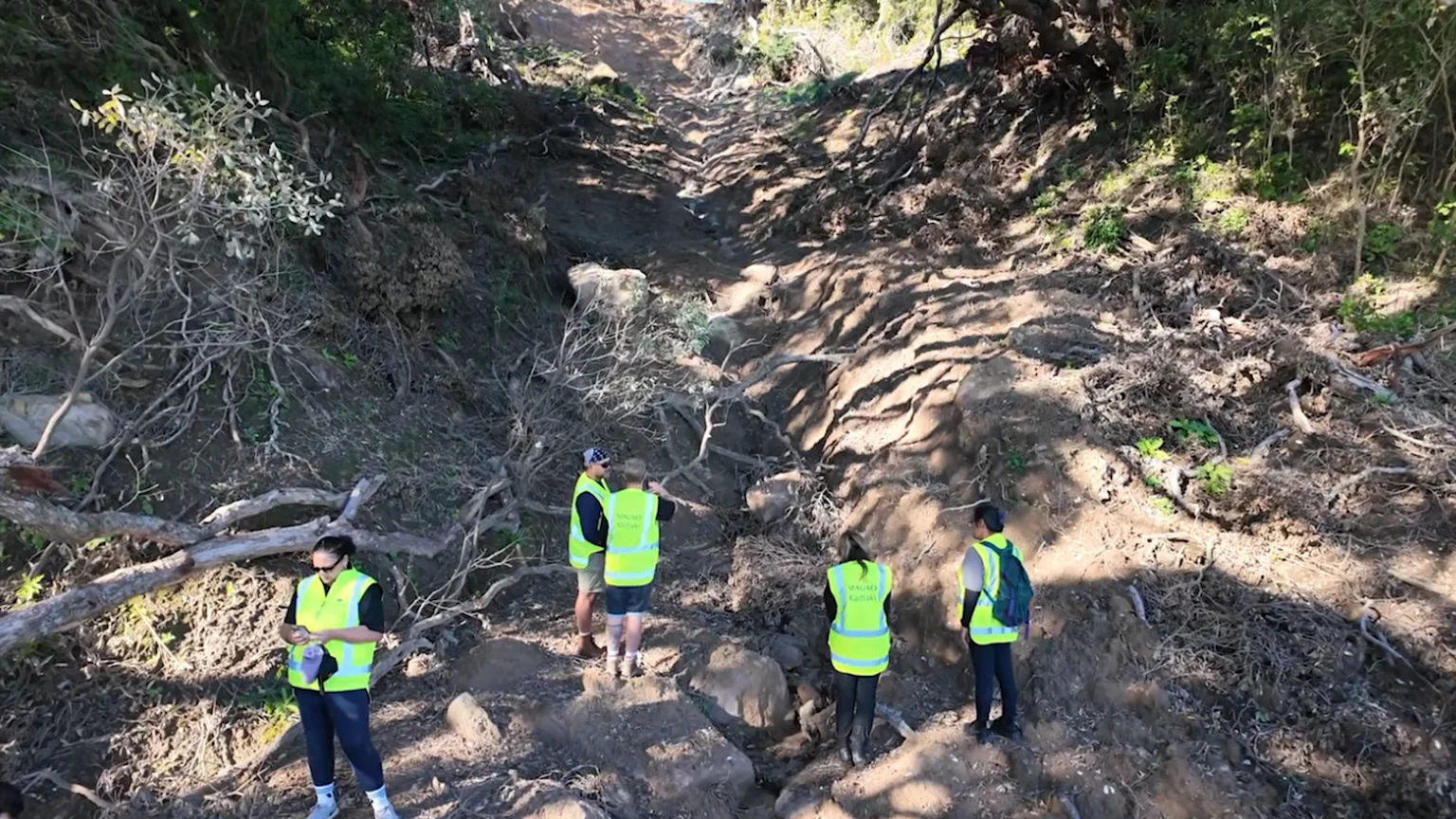 Several people in bright vests stand on an eroded dirt path with steep banks, indicative of a landslide, surrounded by trees and exposed roots.