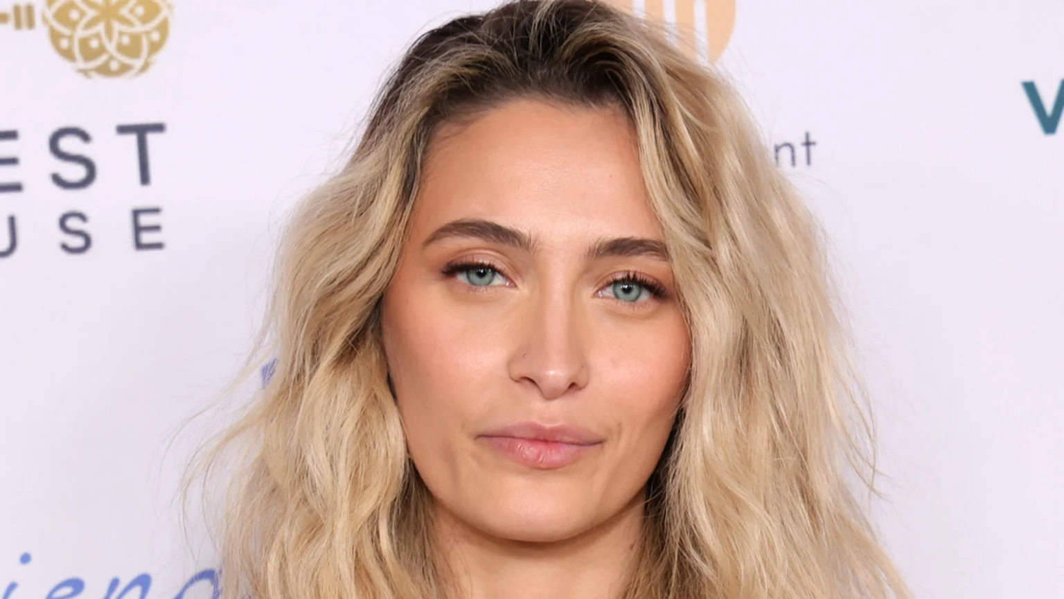 A close-up headshot of Paris Jackson, with wavy blonde hair, light blue eyes, subtle makeup, and a nose piercing, against a blurry white background with logos.