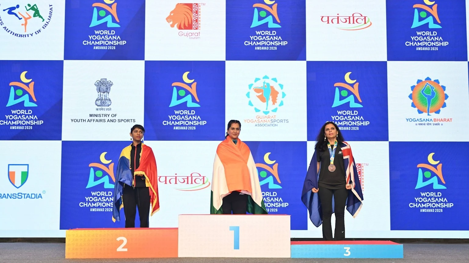 Three women stand on a podium at the World Yogasana Championship: a woman draped in an Indian flag on 1st, another in a yellow, blue, and red flag on 2nd, and a woman wearing a bronze medal and a Union Jack flag on 3rd.