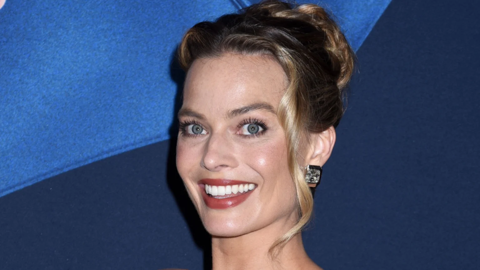 A close-up studio portrait of Margot Robbie smiling directly at the camera. She has blonde hair in an updo, wears a square sparkling stud earring on her right ear, and is set against a bright, diagonally split royal to navy blue background under even lighting.