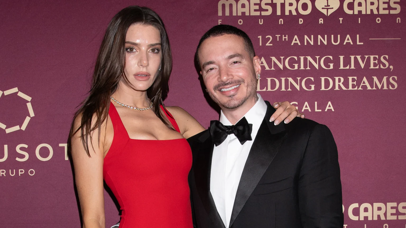 A woman in a red dress and a man in a black tuxedo pose smiling together in front of a burgundy backdrop with gold text, with her hand on his shoulder.