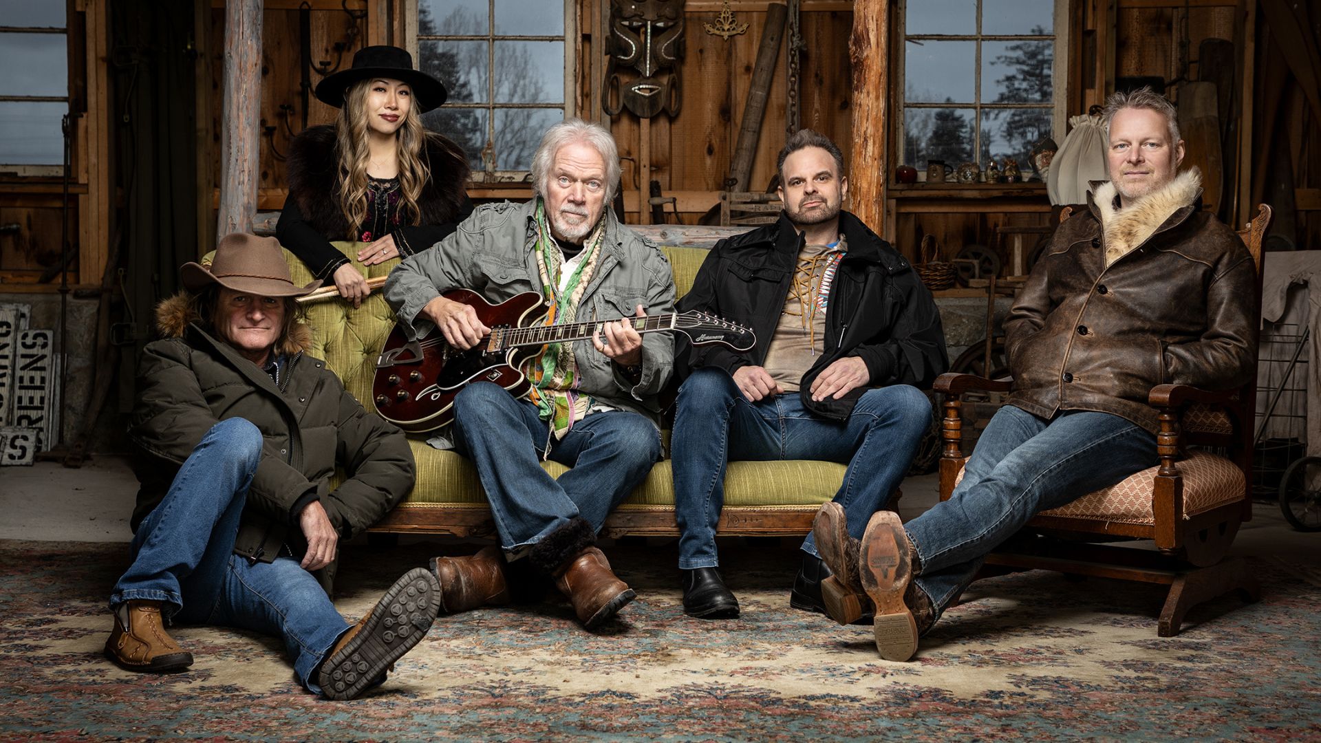 Five people, including a man with a guitar on a couch, a woman standing behind them, and three other men seated, are posed in a rustic, wood-paneled room.