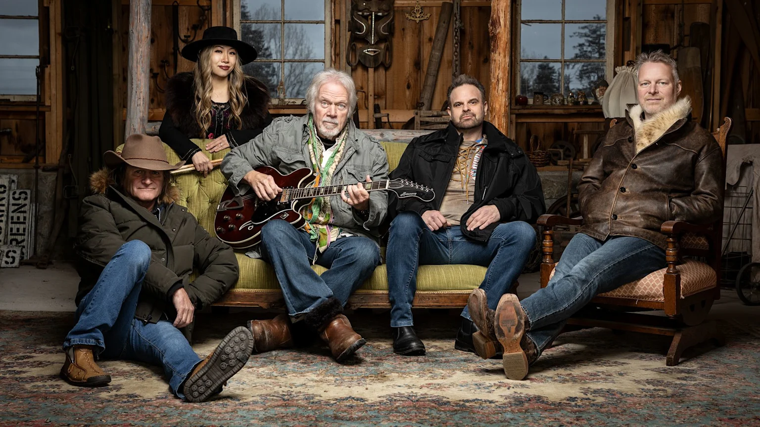 Five people, including a man with a guitar on a couch, a woman standing behind them, and three other men seated, are posed in a rustic, wood-paneled room.