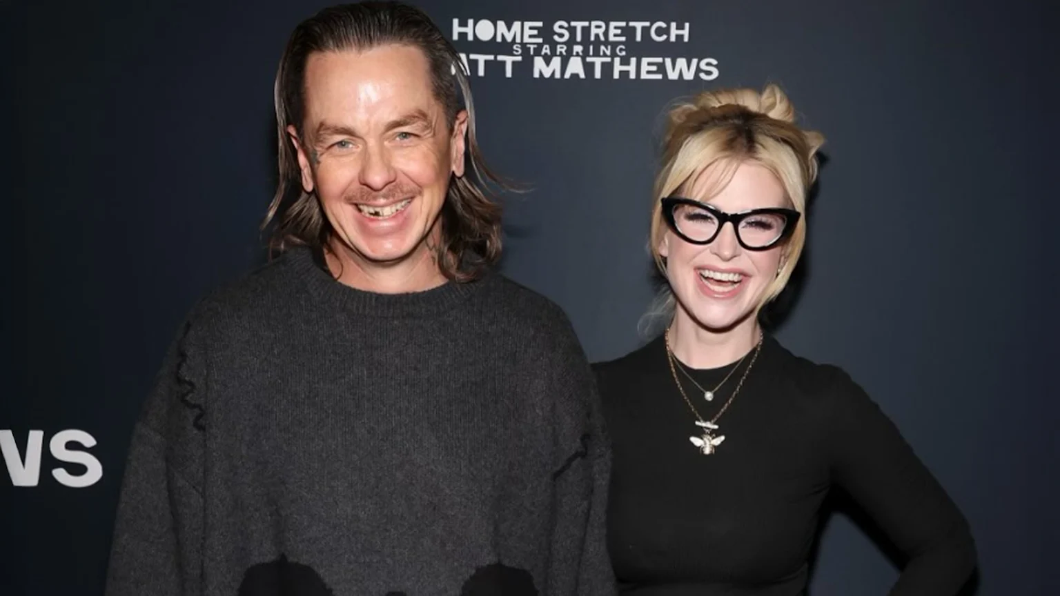 Kelly Osbourne, with blonde hair and cat-eye glasses, laughs beside Sid Wilson, who has long hair, a mustache, and a gap in his front teeth, both smiling for a photo.