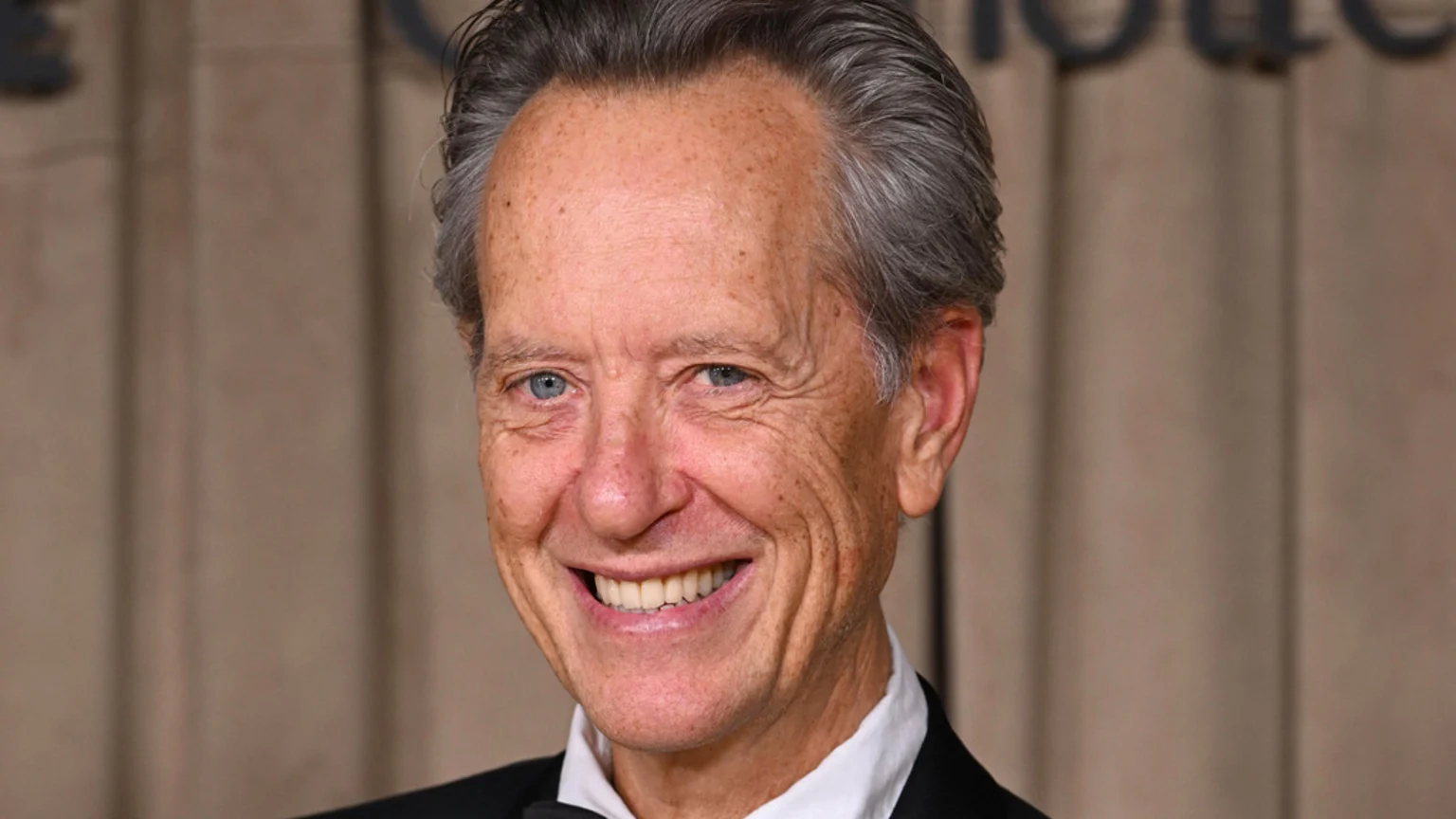 Richard E. Grant, an older man with grey hair and blue eyes, smiles broadly at the camera while wearing a black suit and bow tie.