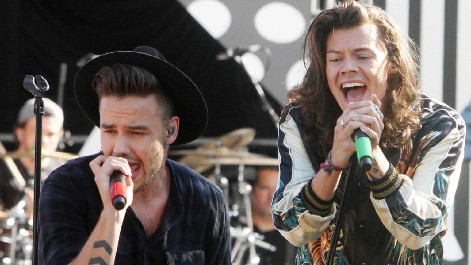 Liam Payne, wearing a dark hat and shirt, sings into a red-tipped microphone on stage alongside Harry Styles, who has long hair, wears a patterned jacket, and sings loudly into a green-tipped microphone.