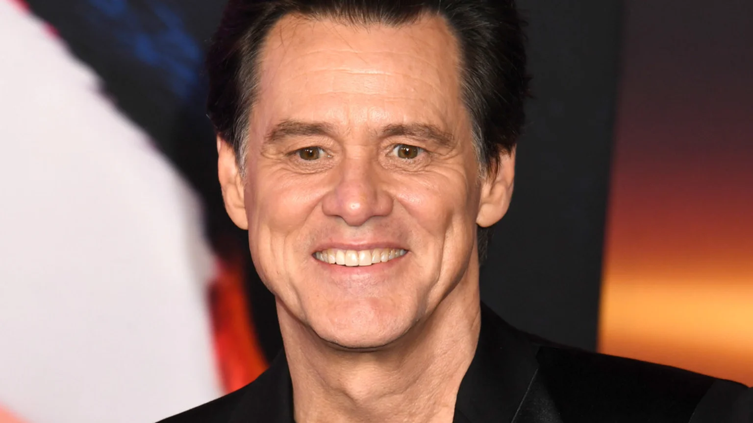 A smiling Jim Carrey in a close-up, looking directly at the viewer, wearing a dark suit jacket against a blurred background with white, dark, and warm orange tones.