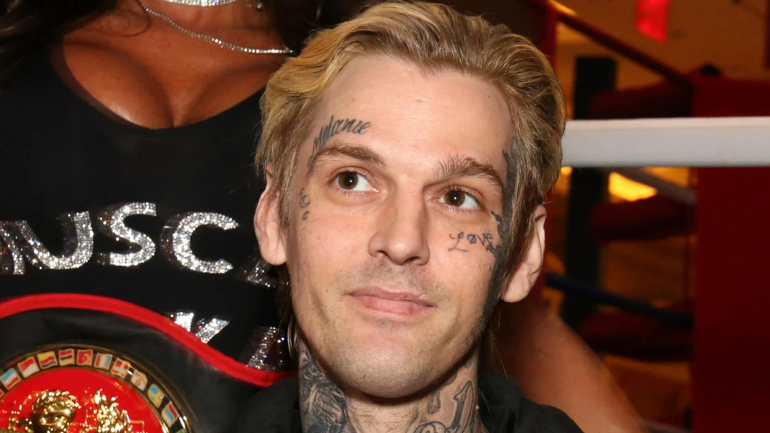 Aaron Carter, a man with blonde hair and face tattoos, looks up and to the right while a woman in a black top with glittery "MUSC" text stands beside him, partly obscuring a boxing belt.