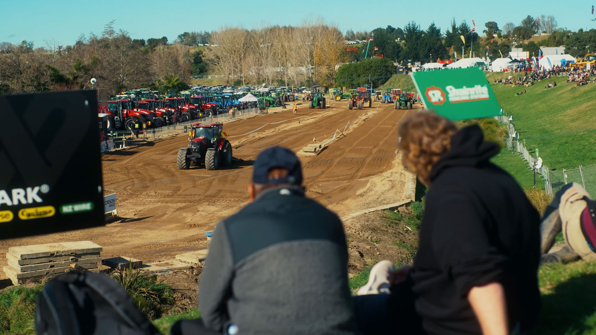 Two spectators watch a tractor pull event on a dirt track, with numerous tractors parked nearby and a crowd of people gathered on a grassy hill under a clear sky.