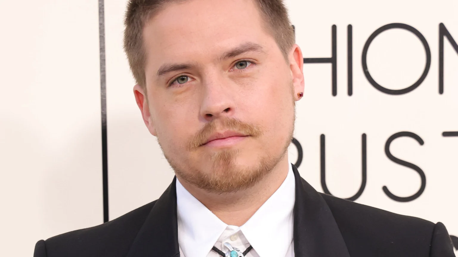 A close-up of a man with light brown hair and a beard, wearing a black suit, white shirt, and a bolo tie with a turquoise stone, and a small red earring in his right ear.