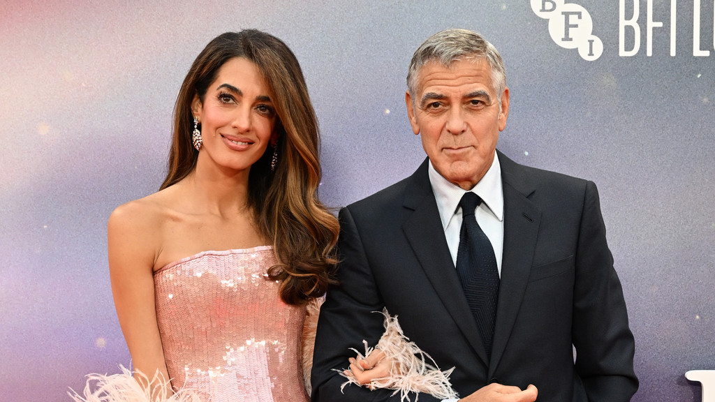 George and Amal Clooney stand side-by-side at an event, with Amal smiling in a pink sequined strapless dress and George looking serious in a dark suit.