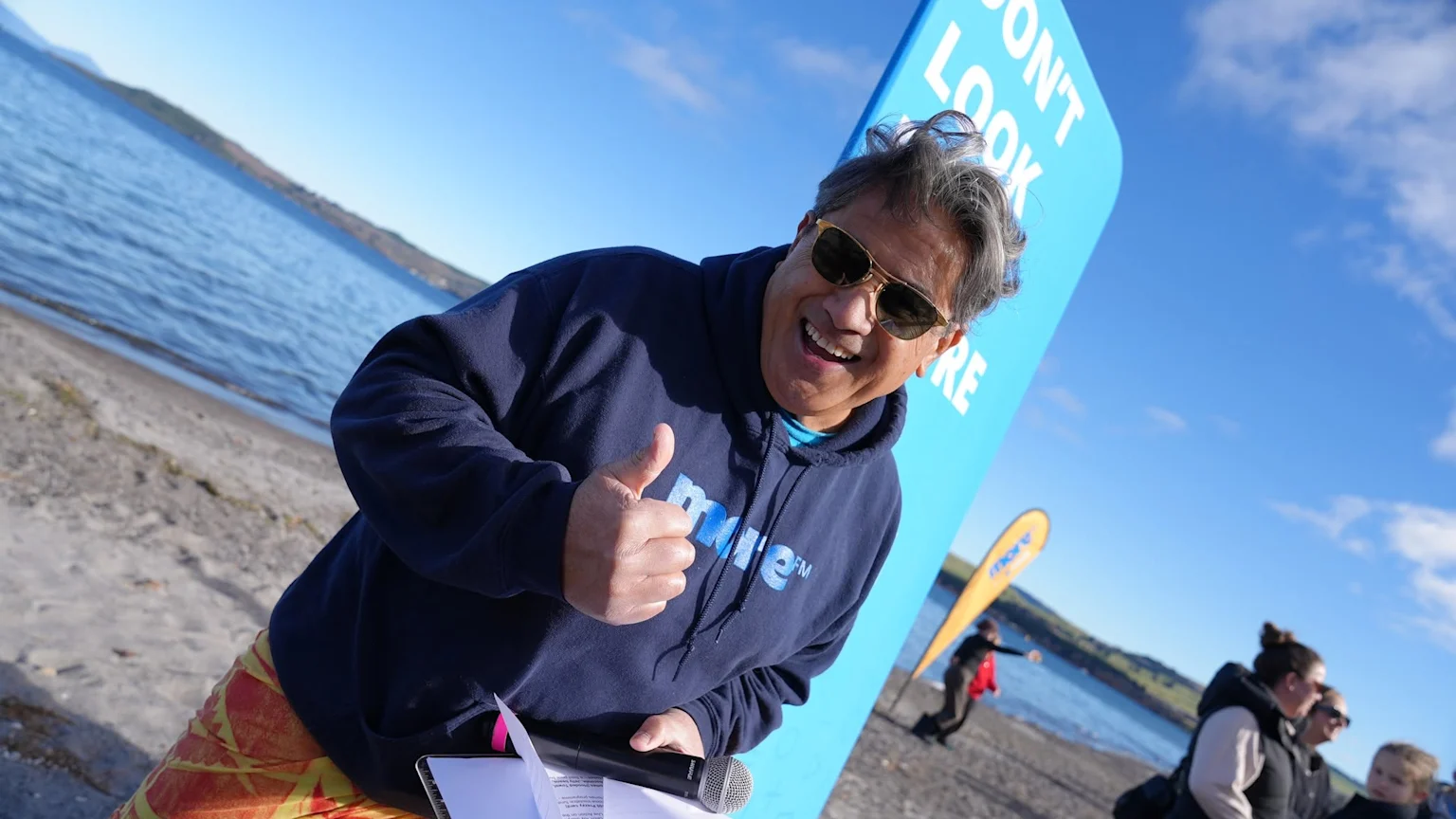 A smiling man in sunglasses, a navy "more FM" hoodie, and orange patterned shorts gives a thumbs up, holding a microphone on a beach with a blue sign and a lake in the background.
