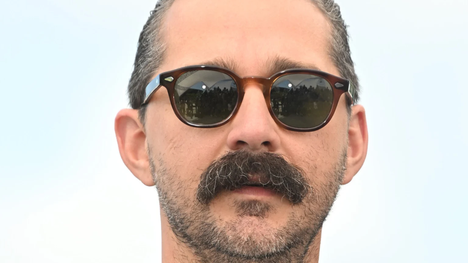 Close-up portrait of Shia LaBeouf wearing round brown sunglasses, a thick dark mustache, and scruffy beard. He wears a light-colored shirt and poses neutrally against a bright clear sky with a blurred crowd and white structures in the background.