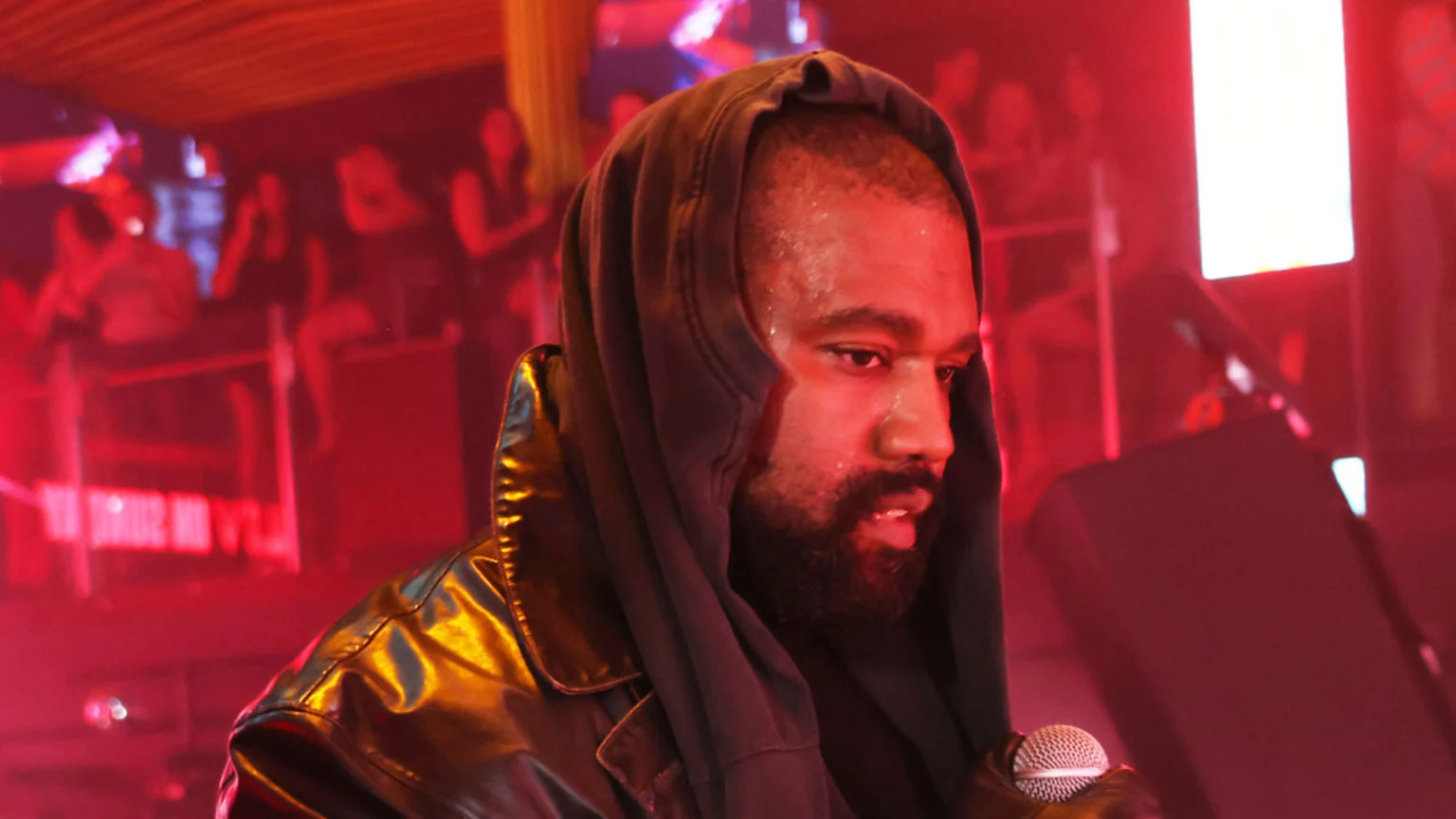 Kanye West, sweating and wearing a dark hoodie over a shiny jacket, holds a microphone under red lighting, looking down intently.