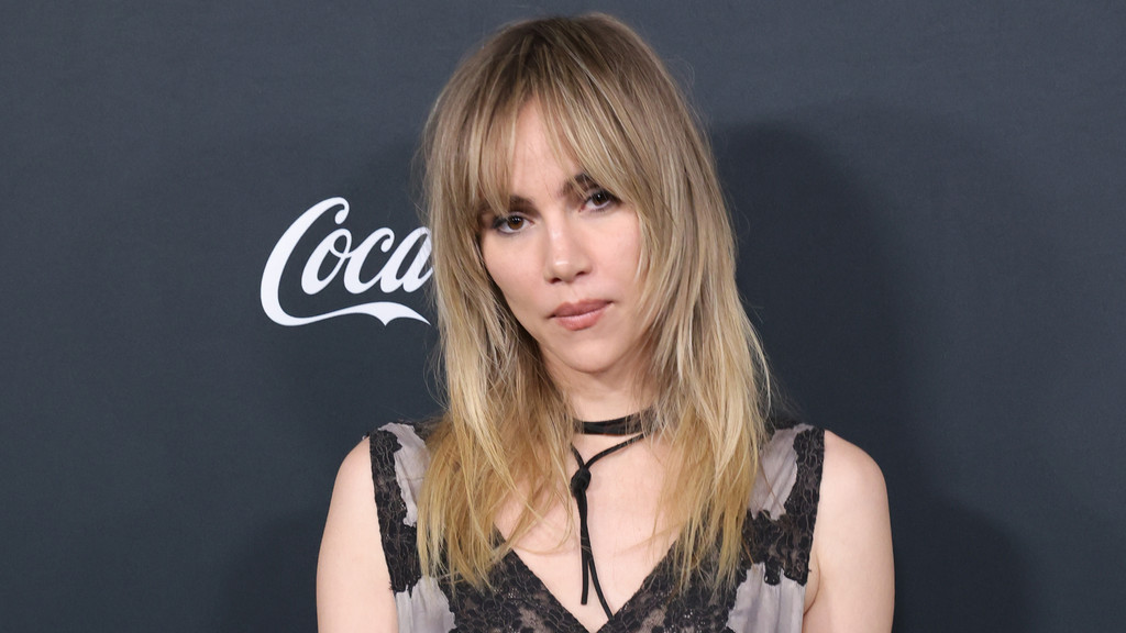 A woman with blonde shaggy hair and bangs, wearing a dark lace top and a choker, looks towards the viewer with a neutral expression against a dark background with a partial Coca-Cola logo.