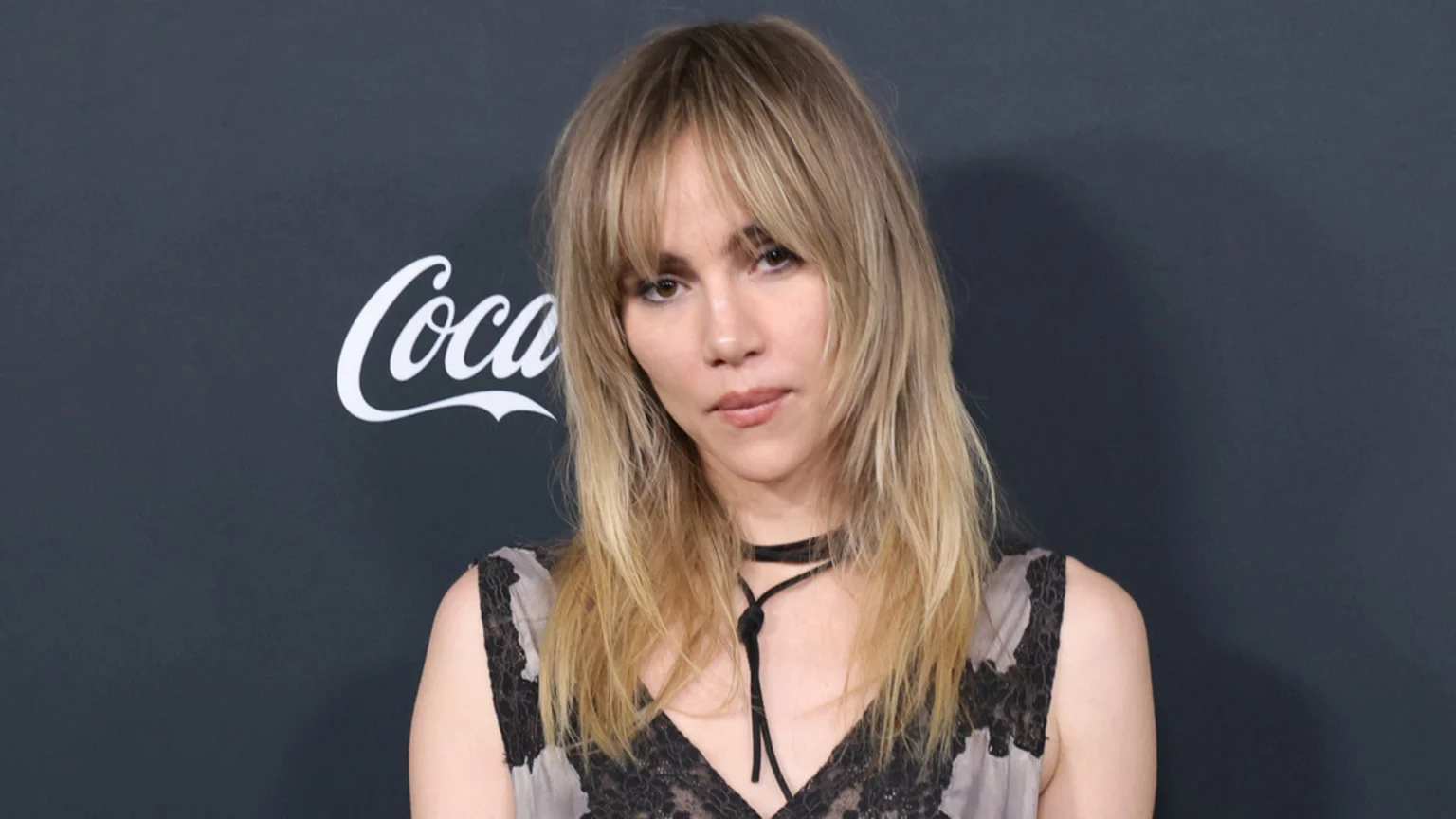 A woman with blonde shaggy hair and bangs, wearing a dark lace top and a choker, looks towards the viewer with a neutral expression against a dark background with a partial Coca-Cola logo.