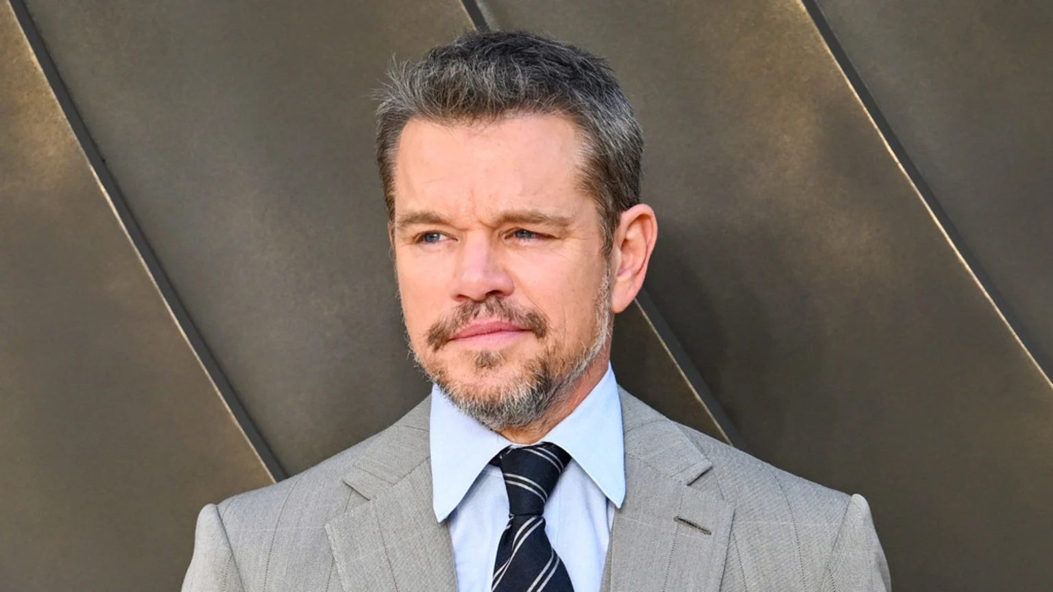 Actor Matt Damon, with salt-and-pepper hair and a beard, looks to the left while wearing a light gray suit, light blue shirt, and dark striped tie, against a dark, diagonally paneled background.