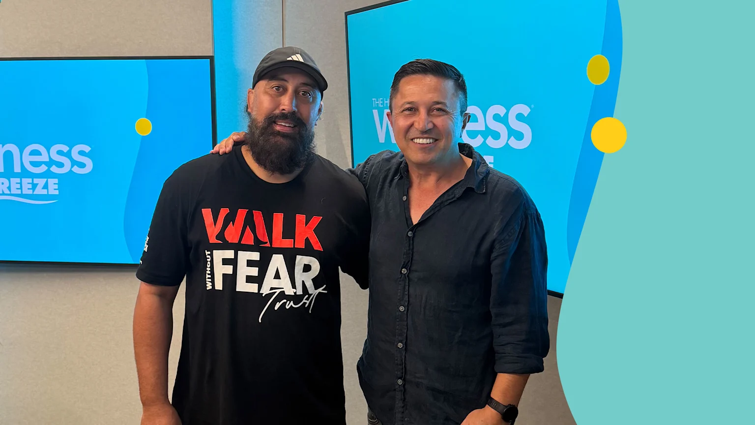 Two men in their 30s or 40s stand close, smiling at the camera. The man on the left wears a black baseball cap and a black t-shirt with the slogan "WALK WITHOUT FEAR Trust." The man on the right wears a dark button-up shirt and wristwatch. Behind them are bright blue screens and abstract teal and yellow graphics.