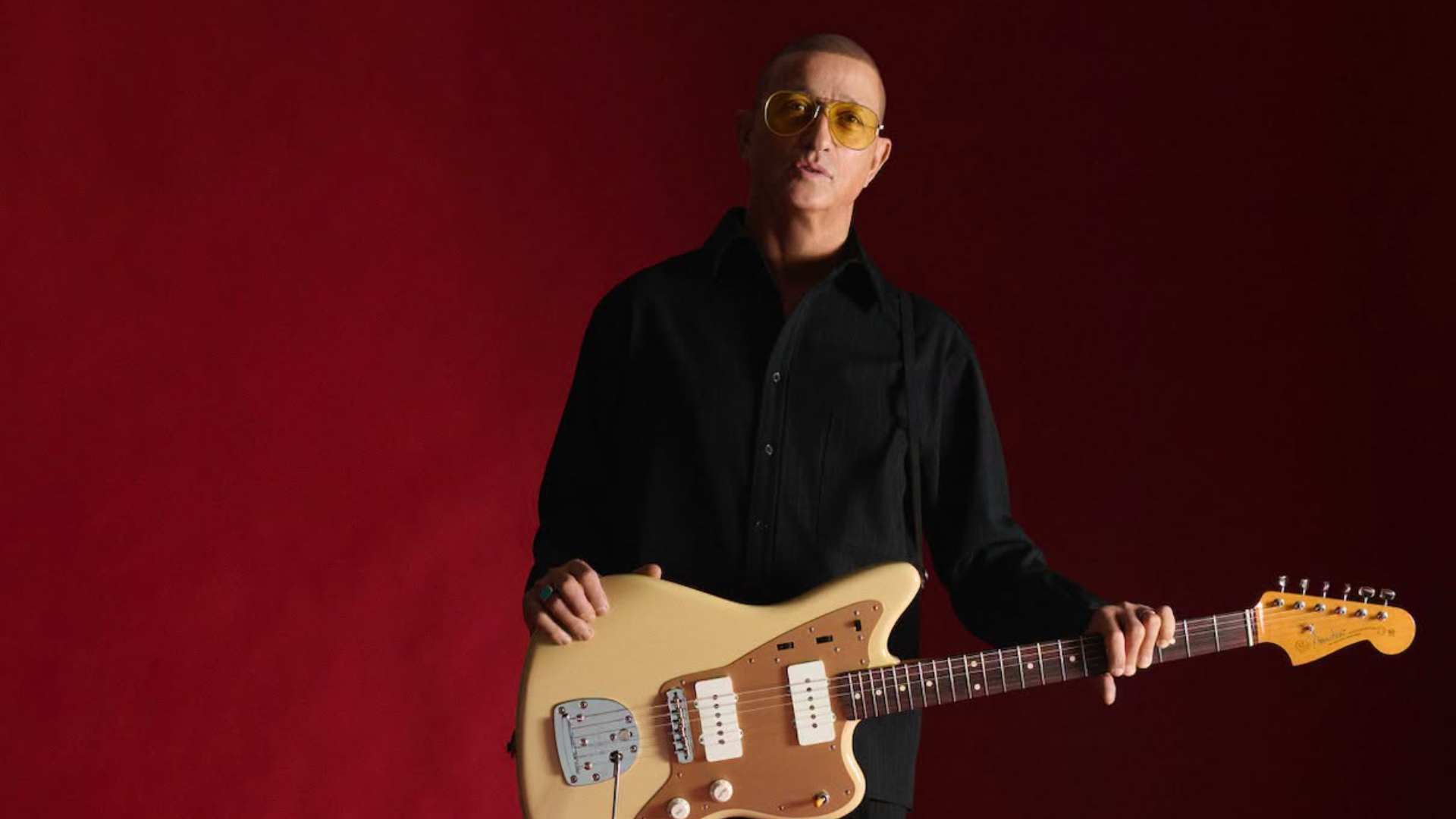 A man with a shaved head and yellow sunglasses, wearing a black shirt, looks at the camera while holding a beige electric guitar against a red background.