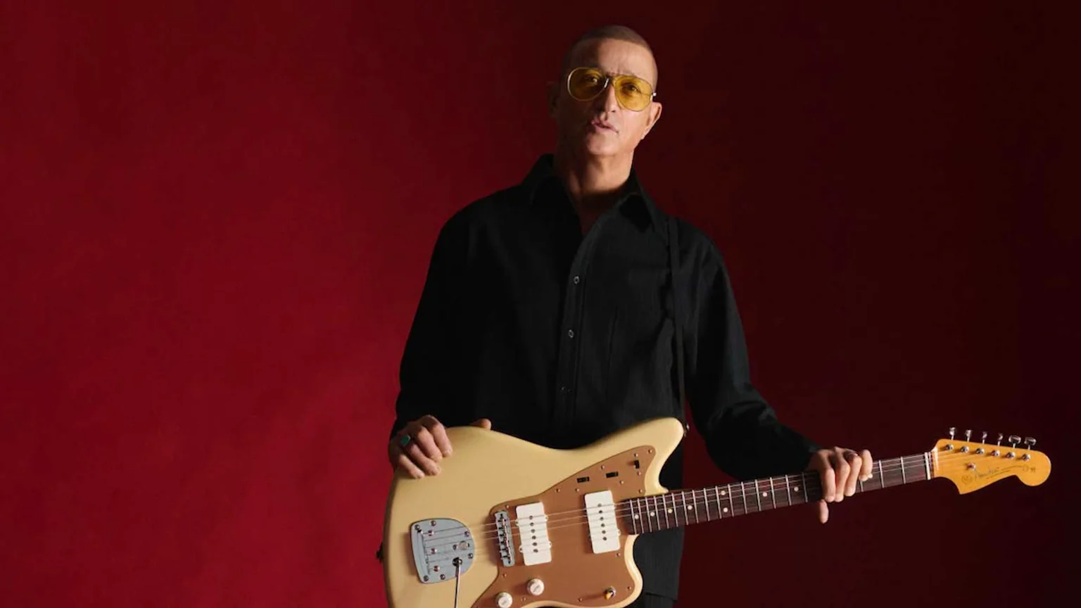A man with a shaved head and yellow sunglasses, wearing a black shirt, looks at the camera while holding a beige electric guitar against a red background.