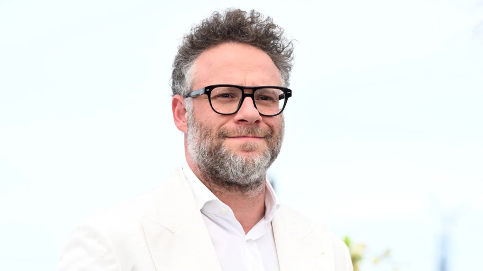 Seth Rogen, wearing black glasses and a white suit, smiles subtly at the camera, showing his curly hair and graying beard.