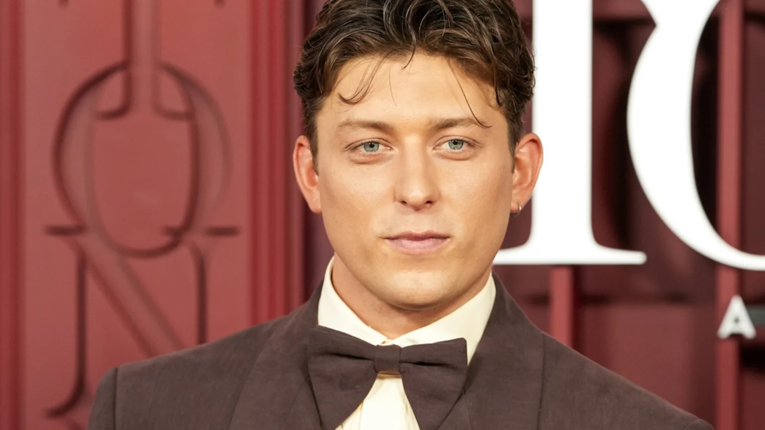 A man with light blue eyes, brown hair, and a small silver hoop earring, wears a brown suit and bow tie, looking directly at the camera against a blurred red background with a large white 'O'.