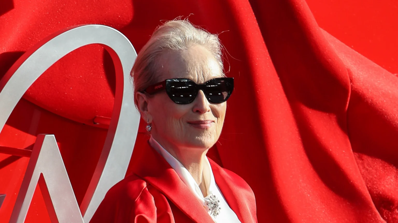 Meryl Streep in a red blazer and white blouse with a brooch, smiling while wearing black Prada sunglasses, against a bright red background with silver accents.