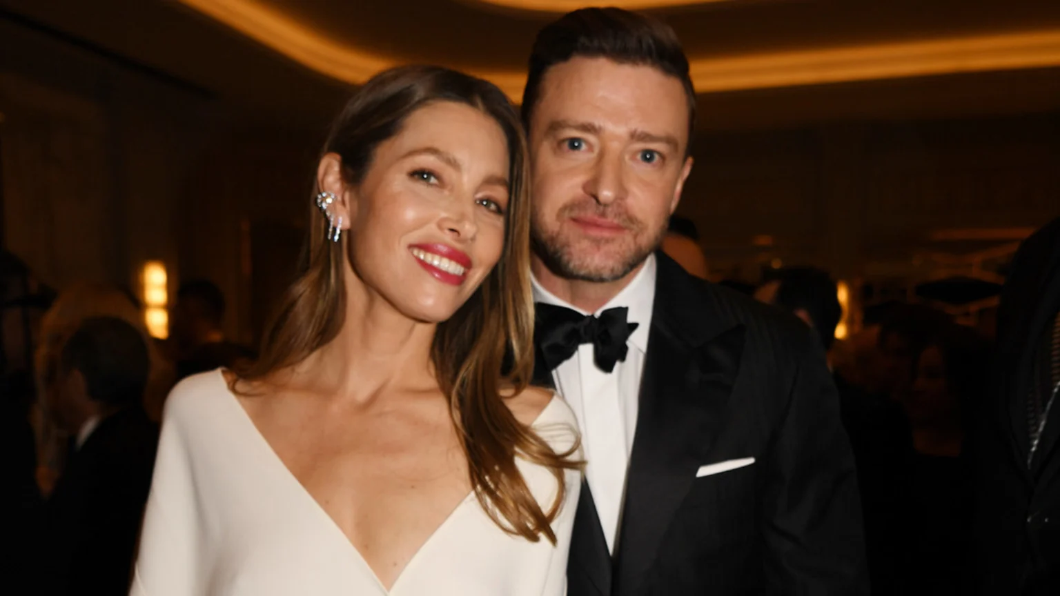 Jessica Biel in a cream V-neck dress and Justin Timberlake in a black tuxedo smile warmly at the camera, standing close together at an event.