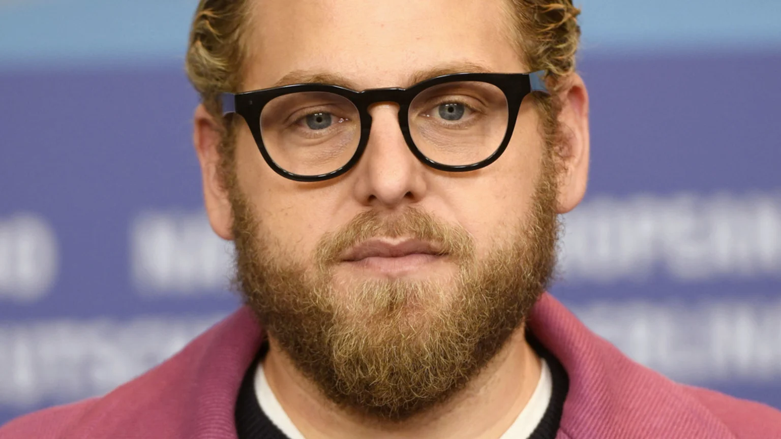 A close-up portrait of actor Jonah Hill looking forward, with light hair, a blonde beard, and black glasses, wearing a pink jacket.