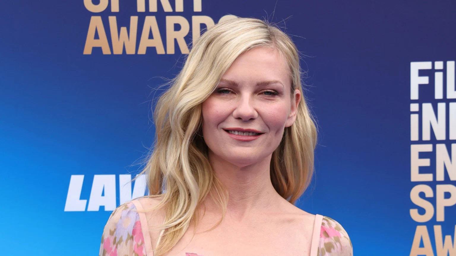 A close-up of actress Kirsten Dunst smiling, with blonde wavy hair, wearing a floral dress, against a blue background with event branding.