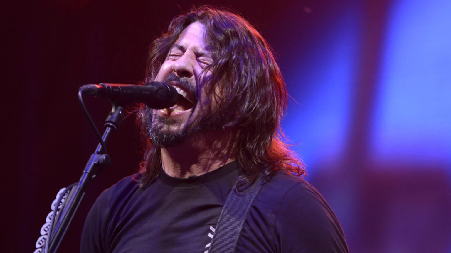 Dave Grohl, with long dark hair and a beard, intensely sings into a microphone on stage, eyes closed and mouth open, under purple and blue stage lights.