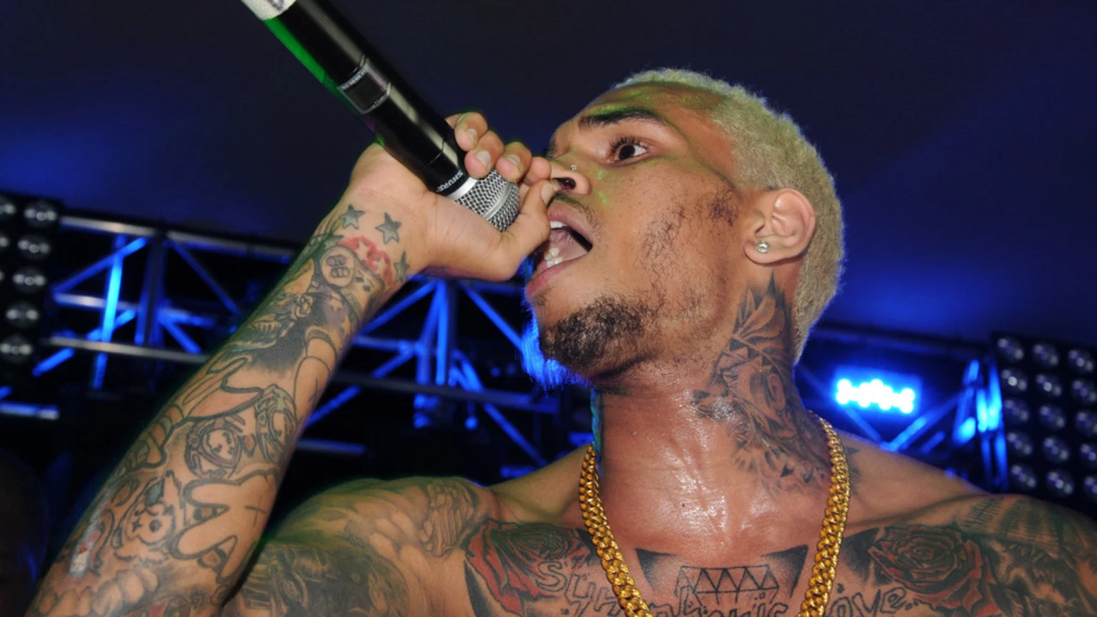 A sweaty Chris Brown, with blonde hair, a beard, and extensive tattoos, sings into a microphone on stage, wearing a gold chain and an earring, with blue stage lights in the background.