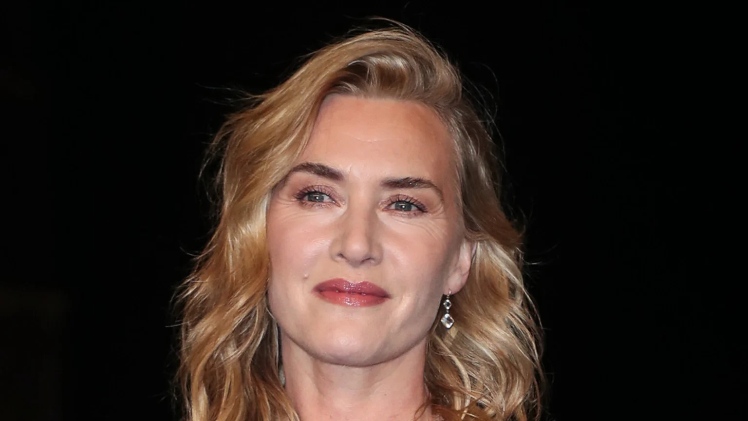 A close-up of actress Kate Winslet, with blonde wavy hair, blue eyes, and light pink lipstick, smiling subtly against a dark background.